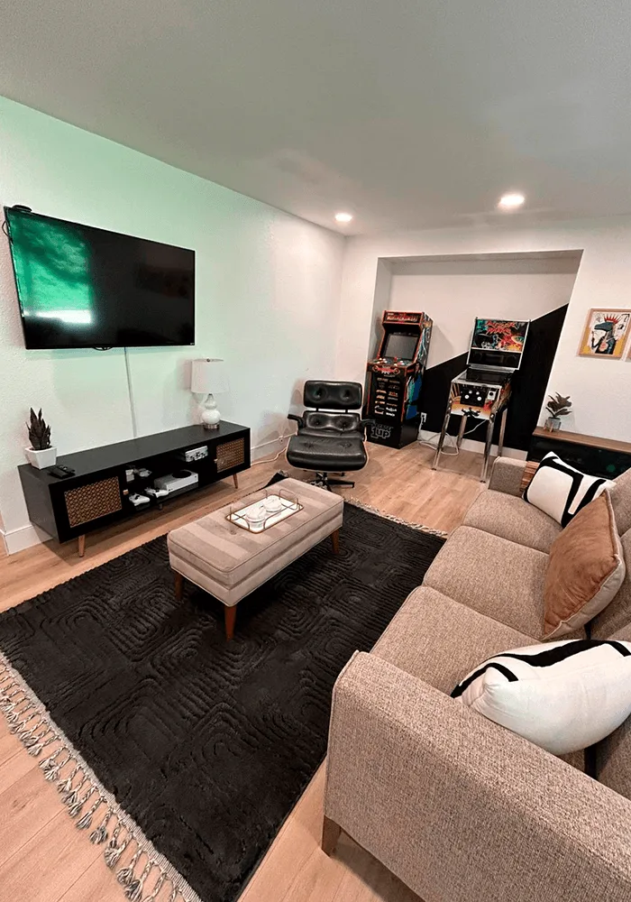 A clean and tidy game room with a sectional sofa, a large TV, arcade machines, and a professionally cleaned area rug over laminate flooring.